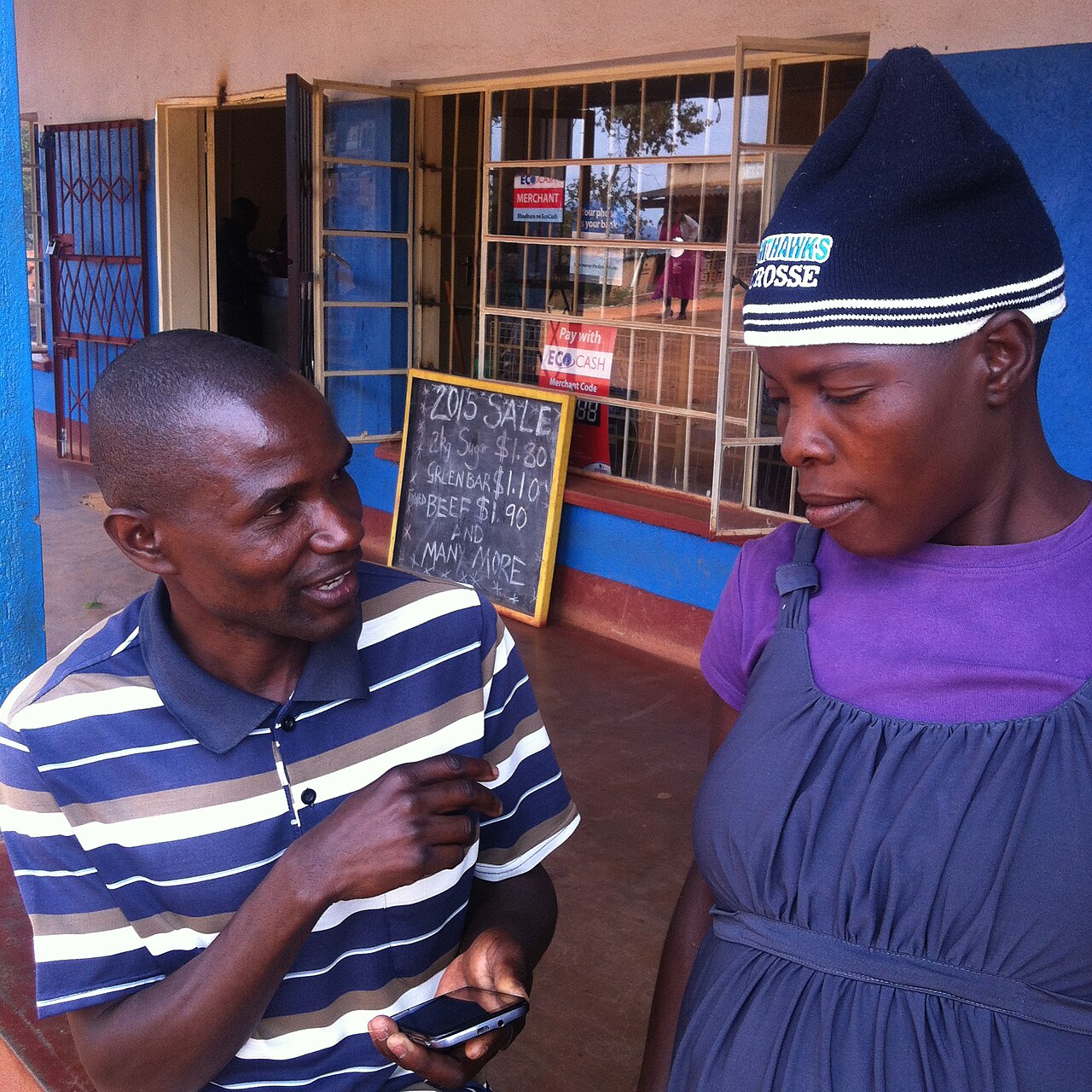 A survey enumerator holding a mobile phone interviews a woman outside a village shop in Mutasa, Zimbabwe