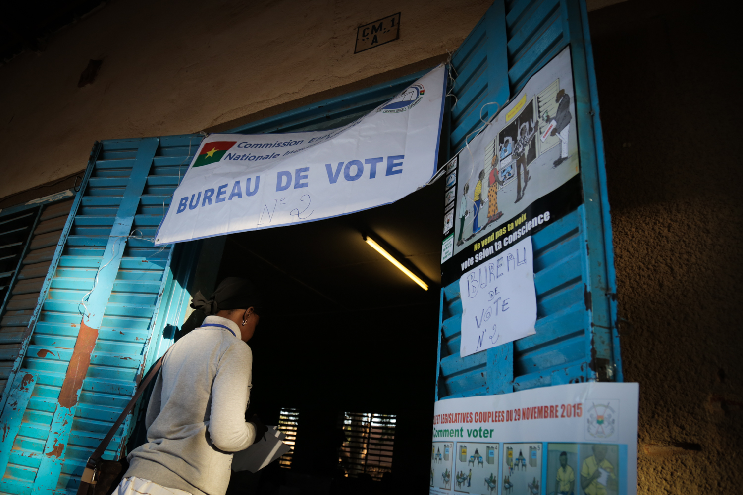 A voter enters a polling station with blue shutters in Ouagadougou. Banners read Bureau de vote N° 2, and a poster says Ne vend pas ta voix, vote selon ta conscience.