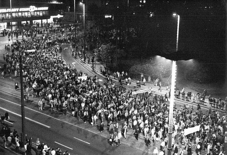 Night view from above of a dense crowd marching along a wide, lamp-lit street in Leipzig
