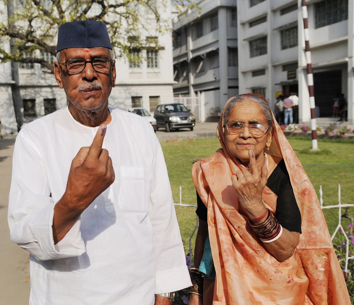 An elderly couple, he in a white kurta and cap, she in an orange sari, hold up ink-marked fingers outside a polling station in Patna