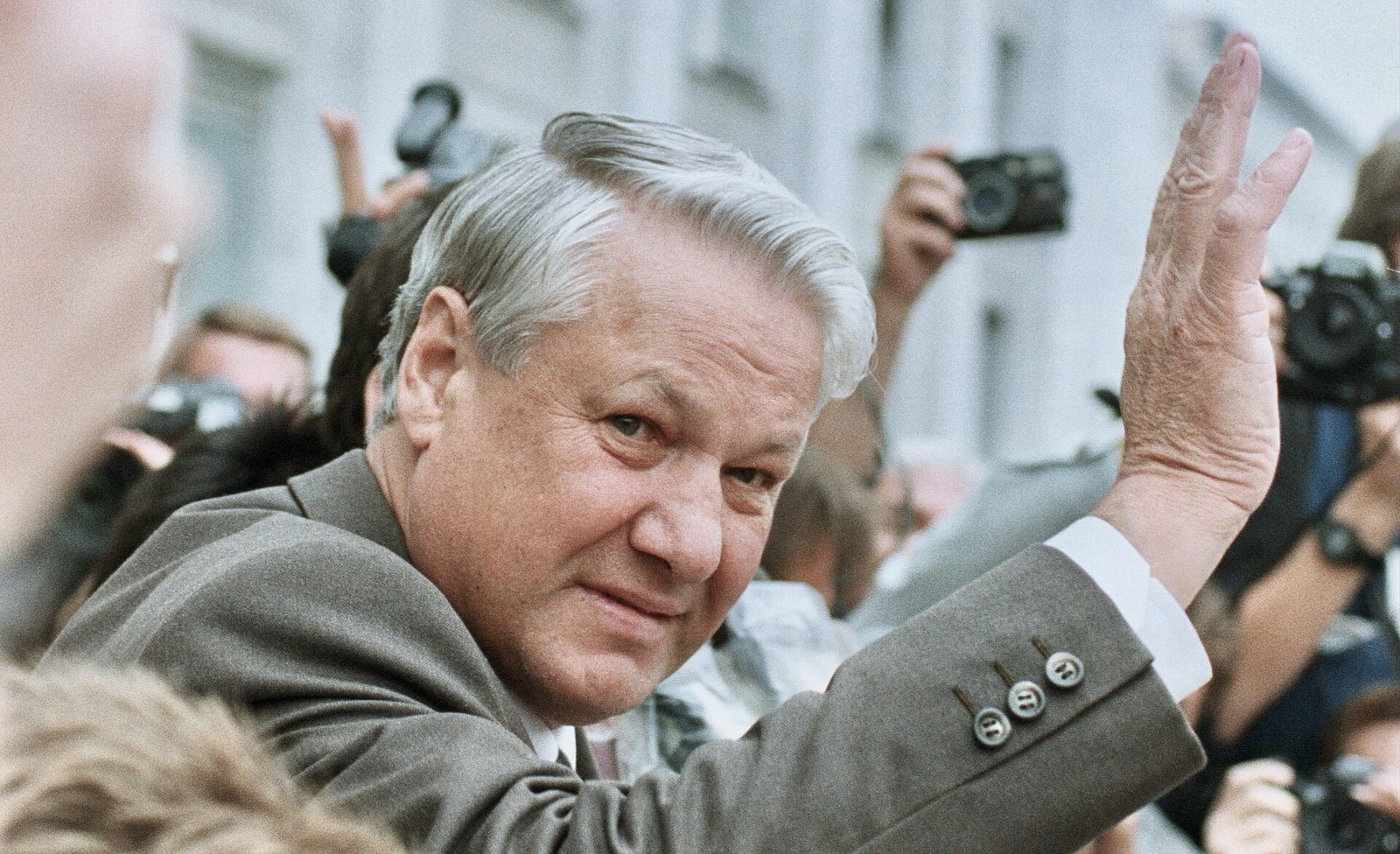 Boris Yeltsin, smiling, raises his hand to a crowd of photographers holding up cameras
