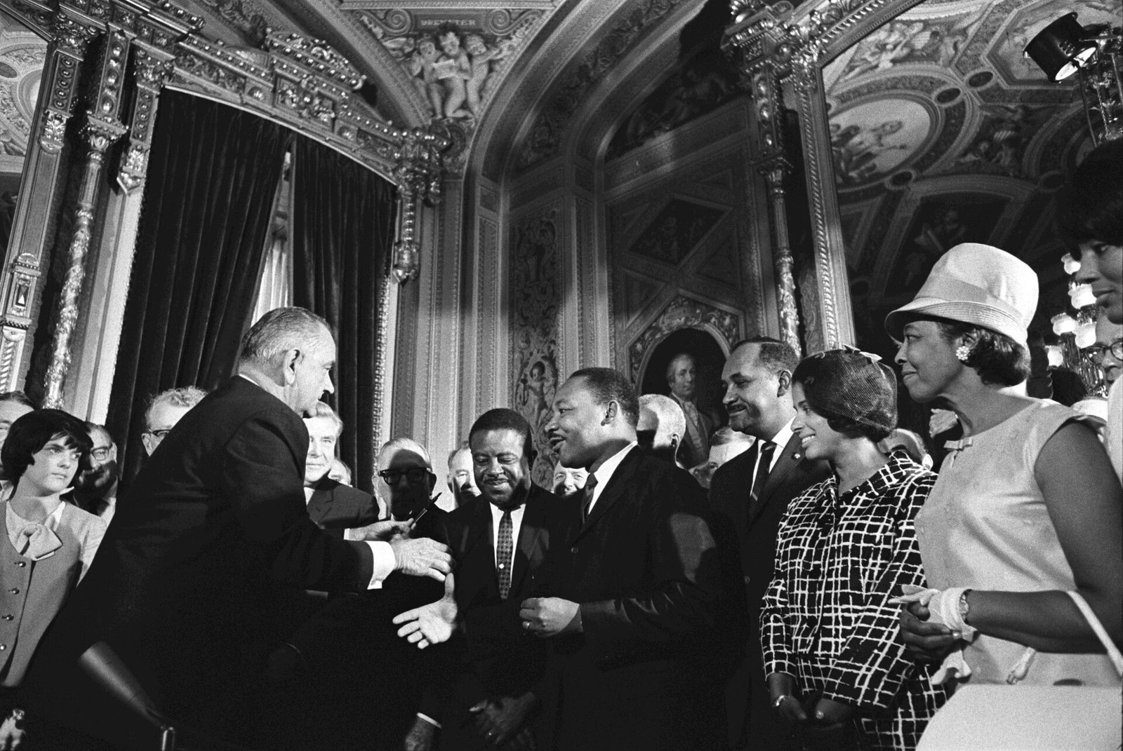 President Lyndon B. Johnson reaches to shake hands with Martin Luther King Jr. in an ornate room of the Capitol, with civil rights leaders around them